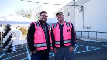 two men in pink vests standing in a parking lot smiling for the camera