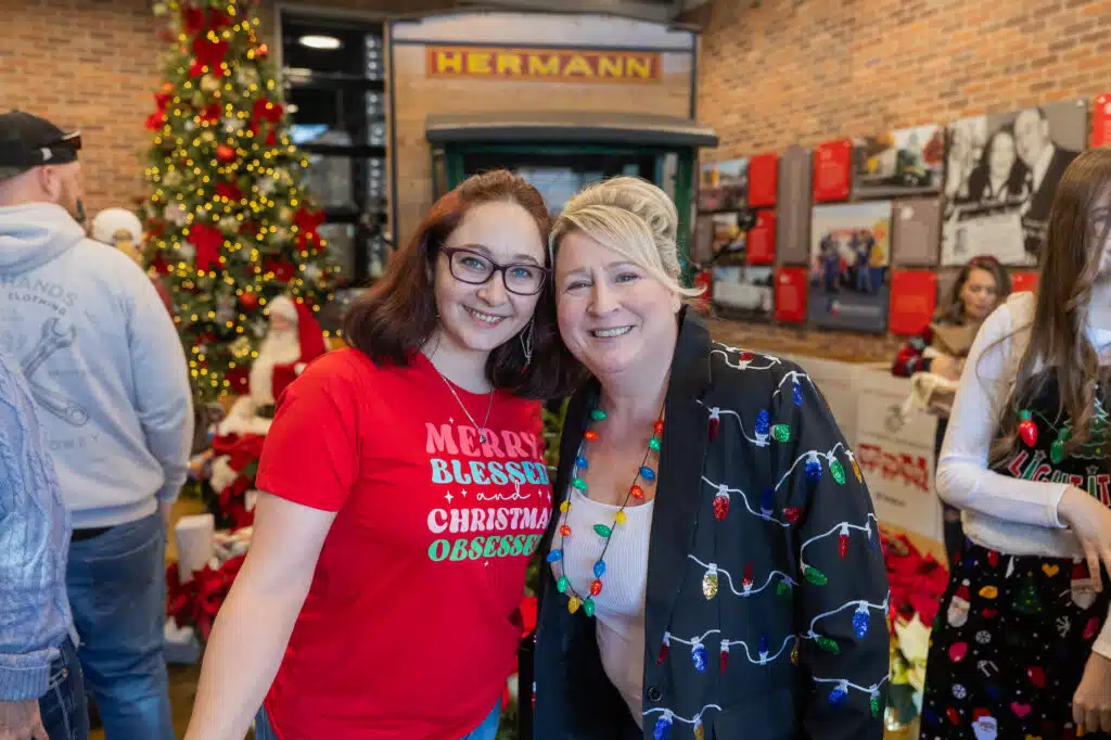 two smiling women stand side by side in front of an old green truck surrounded by toy donation boxes and Christmas decorations